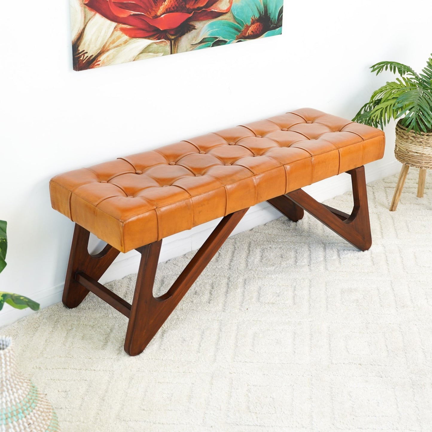 The Mia Tufted Leather Wood Base Bench 47 by Ashcroft Furniture Co. has genuine leather upholstery and wood legs, styled on a light rug near a potted plant and colorful floral art for a Mid-Century Modern vibe.
