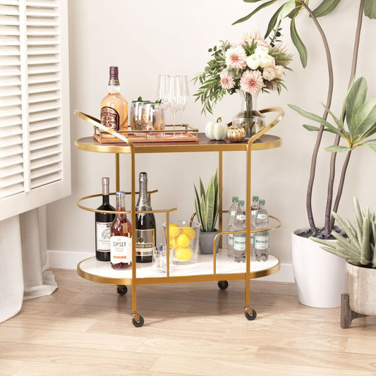 The ZUO Modern Antalya Swanky Two Tier Bar Cart, in gold, holds liquor bottles, glasses, mixers, a lemon tray, and a flower vase beside potted plants and a white shuttered window in a bright, cozy room.