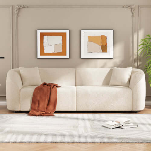 Contemporary sloped arm boucle upholstered sofa in cream with orange throw, styled in a modern living room.