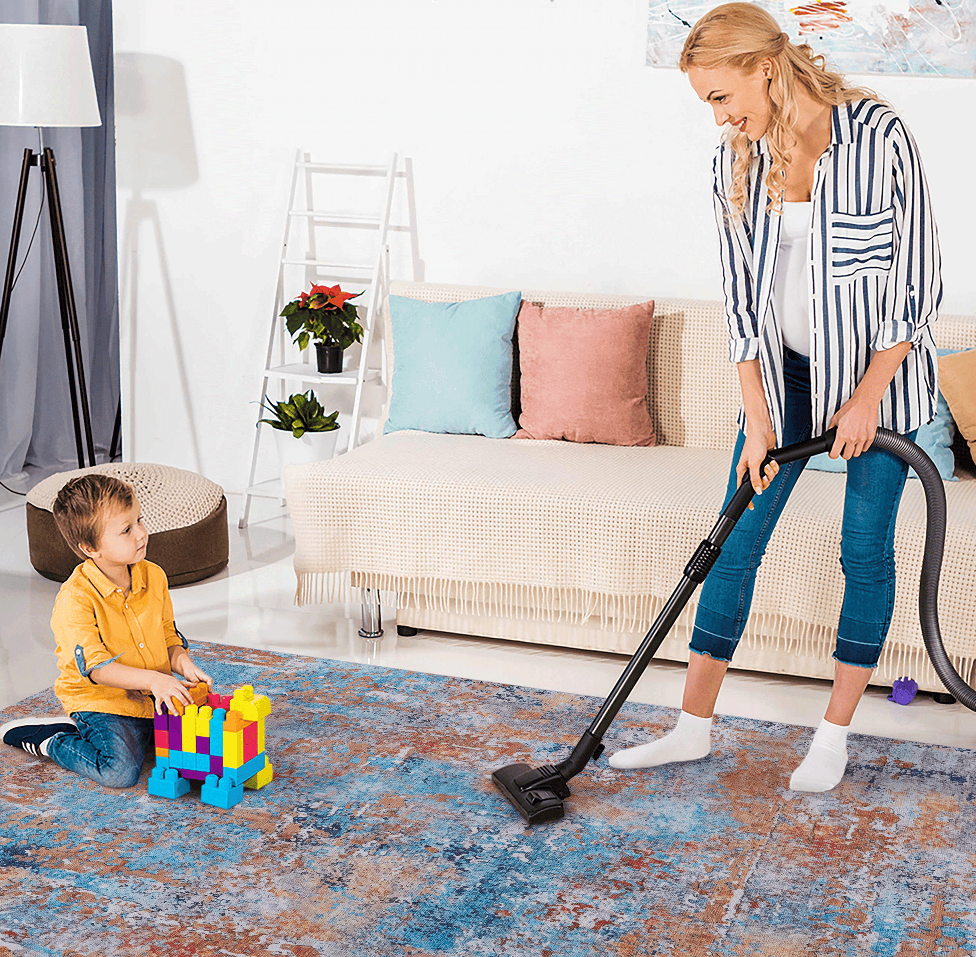 A woman vacuuming a colorful abstract area rug while a child plays with building blocks nearby.