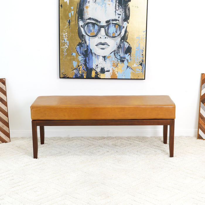 The Austin MCM Genuine Leather Bench 51 by Ashcroft Furniture with wooden legs sits on a white textured rug below a modern painting of a woman in sunglasses, set against an abstract blue and gold-accented wall.