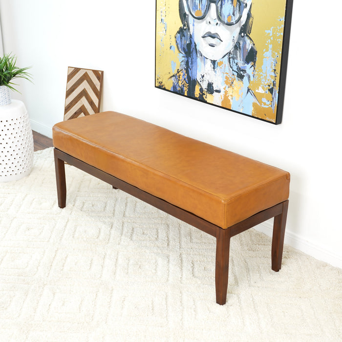 The Ashcroft Furniture Austin MCM Genuine Leather Bench 51 with dark wood legs rests on a white textured rug in a modern room, accented by a colorful portrait, chevron-patterned decor, and a potted plant for Mid-Century Modern flair.