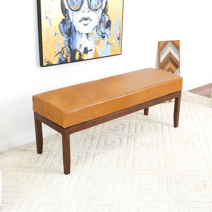 The Ashcroft Furniture Austin MCM Genuine Leather Bench 51, featuring caramel-colored leather and wooden legs, sits on a cream patterned rug. Above it hangs a colorful painting of a womans face with sunglasses.