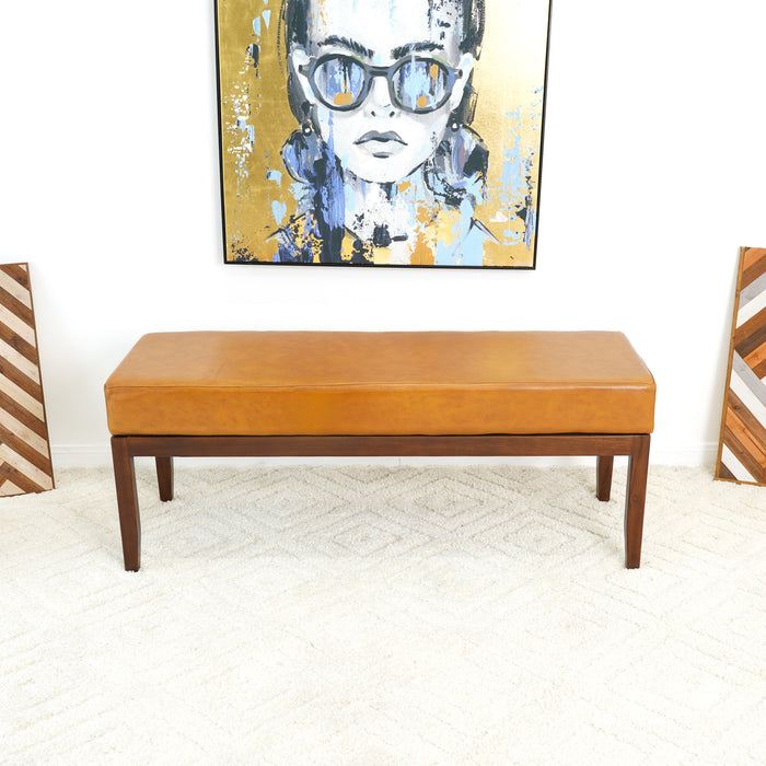 A brown Ashcroft Furniture Austin MCM Genuine Leather Bench 51 with wooden legs sits on a light textured rug beneath a colorful abstract portrait of a woman with glasses. Two chevron-patterned panels lean against the side walls.