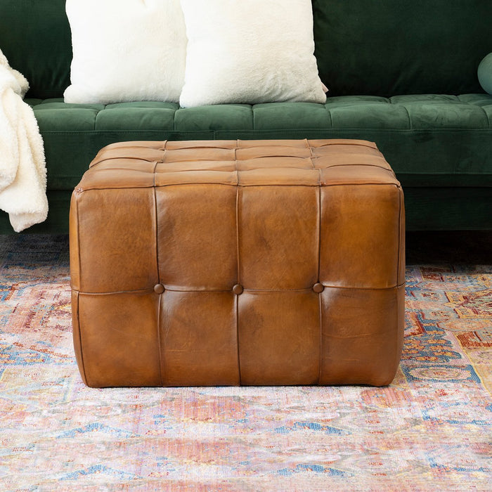 The Benjiro Button Tufted Square Leather Ottoman by Ashcroft Furniture Co sits on a colorful rug in front of a green velvet sofa with white pillows and a white blanket.