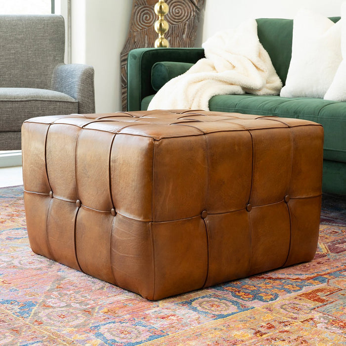 The Benjiro Button Tufted Square Leather Ottoman 27 by Ashcroft Furniture Co sits on a colorful rug in a cozy living room, paired with a green sofa, white blanket, and gray armchair in the background.