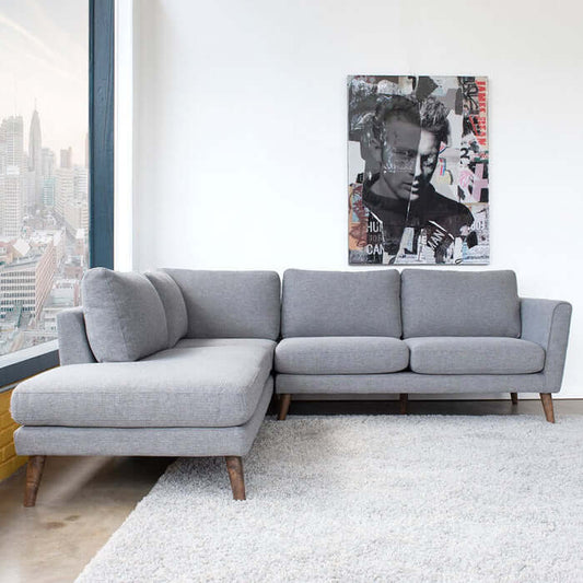 A modern living room with the Ashcroft Furniture Co. Benson Fabric L Shape Sectional Chaise Sofa 97 on a white rug, large window with city view, and black-and-white portrait artwork on the wall.