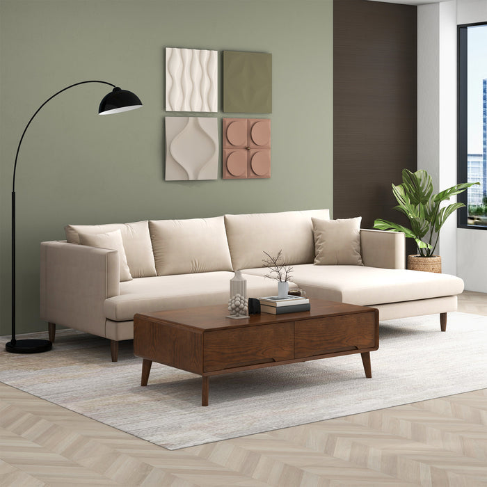A modern living room with the Blake Velvet Upholstered L-Shape Chaise Sofa Sectional 107 by Ashcroft Furniture in cream, a wood coffee table, black floor lamp, green accent wall with geometric art, potted plant, and city views through large windows.