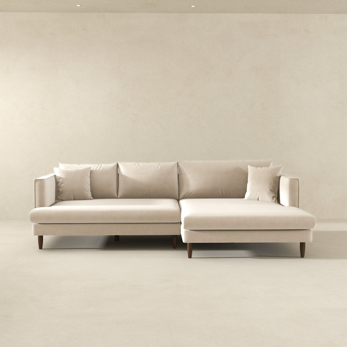 The Blake Velvet Upholstered L-Shape Chaise Sofa Sectional 107 by Ashcroft Furniture, with plush velvet, reversible chaise, four back cushions, and two accent pillows, is set in a minimalist room with beige walls and floor.