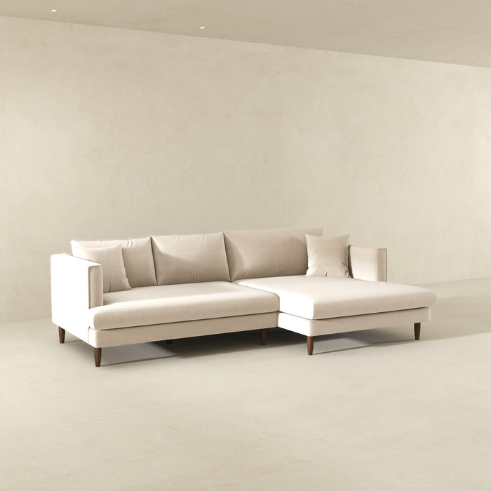 The Ashcroft Furniture Blake Velvet Upholstered L-Shape Chaise Sofa Sectional 107 in light beige, featuring plush velvet, wooden legs, three back cushions, two throw pillows, and a reversible chaise, adds modern style to a minimalist room with off-white walls and floor.