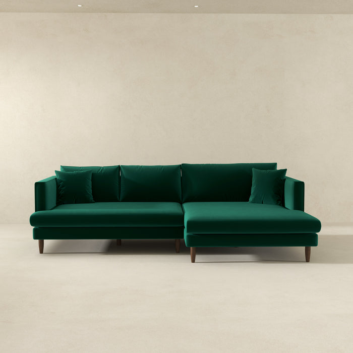 The Blake Velvet Upholstered L-Shape Chaise Sofa Sectional 107 by Ashcroft Furniture, with plush velvet upholstery, wooden legs, and matching cushions, adds elegance to a minimalist room with beige walls and a light floor.