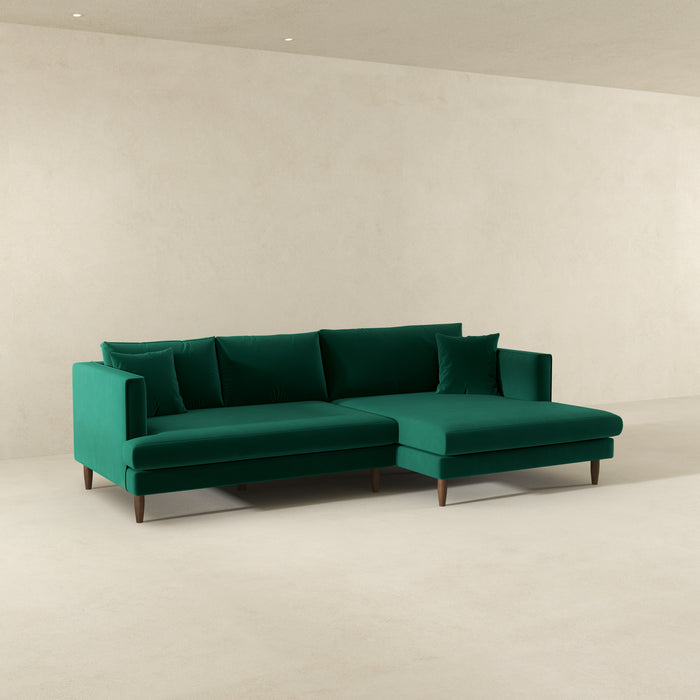 The Ashcroft Furniture Blake Velvet Upholstered L-Shape Chaise Sofa Sectional 107 features plush velvet upholstery, clean lines, and a reversible chaise, shown in a minimalist room with plain beige walls and floor.