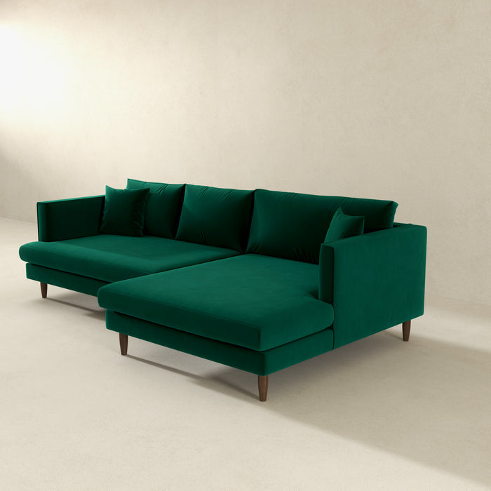 The Blake Velvet Upholstered L-Shape Chaise Sofa Sectional 107 by Ashcroft Furniture, featuring plush velvet and wooden legs, is set in a minimalist light room with four matching green cushions.