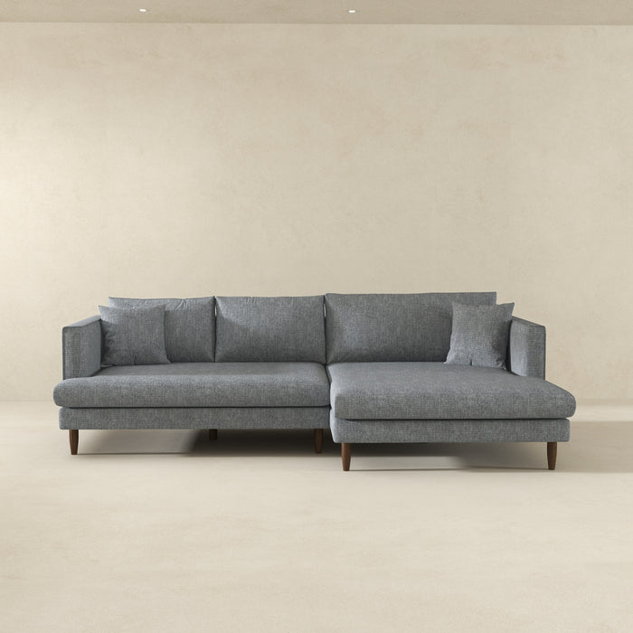 The Ashcroft Furniture Blake Velvet Upholstered L-Shape Chaise Sofa Sectional 107 in modern gray stands against a plain light beige wall and floor, adding comfort and style to a minimal, spacious room.
