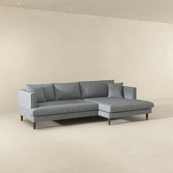The Ashcroft Furniture Blake Velvet Upholstered L-Shape Chaise Sofa Sectional 107, in modern gray, featuring plush velvet and wooden legs, is placed in a minimalist room with light beige walls and floors.