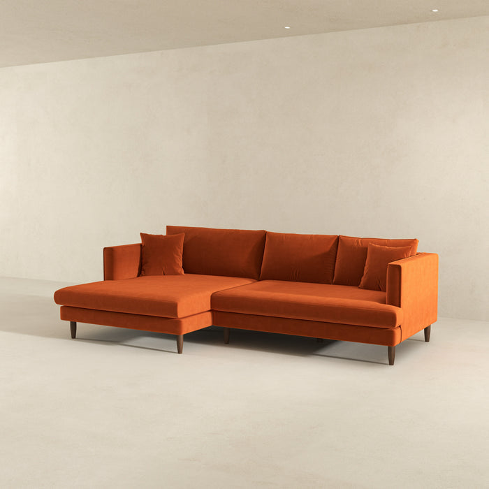 The Blake Velvet Upholstered L-Shape Chaise Sofa Sectional 107 by Ashcroft Furniture, featuring plush velvet and wooden legs, sits in a minimalist, light-colored room with plain walls and no other furnishings.