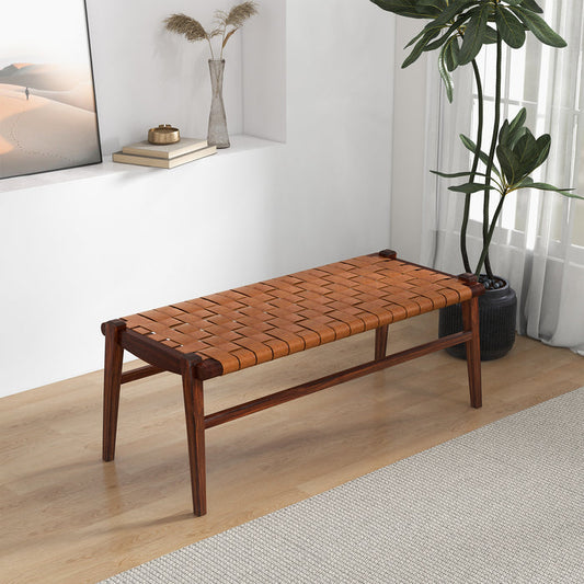 The Cody MCM Strapped Genuine Leather Bench 44 by Ashcroft Furniture, featuring a tan woven leather seat, stands on a light wood floor near a plant in a bright, mid-century modern room with white walls and built-in shelving.