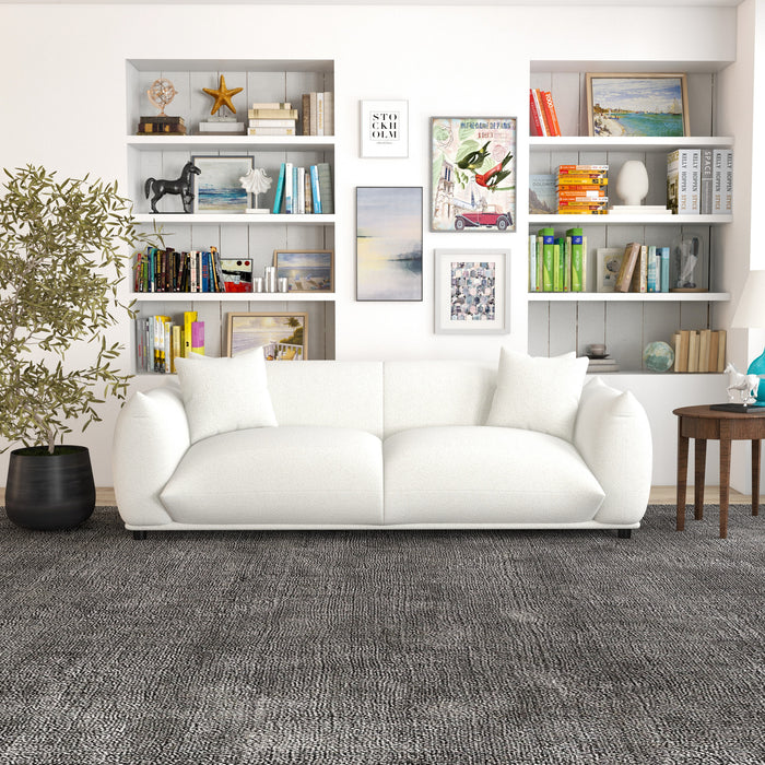 A modern living room features the Ashcroft Furniture Emma Boho Cream Boucle Upholstered Sofa 89, two white pillows, a gray textured rug, a round wooden side table, a potted plant, and wall shelves with books, art, and decor for Bohemian flair.
