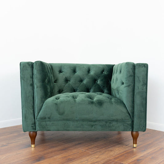 The Ashcroft Furniture Evelyn Mid-Century Tufted Velvet Lounge Chair 42 features plush green velvet, high sides, and wooden legs with gold tips, embodying classic mid-century style against a plain white wall on a wooden floor.