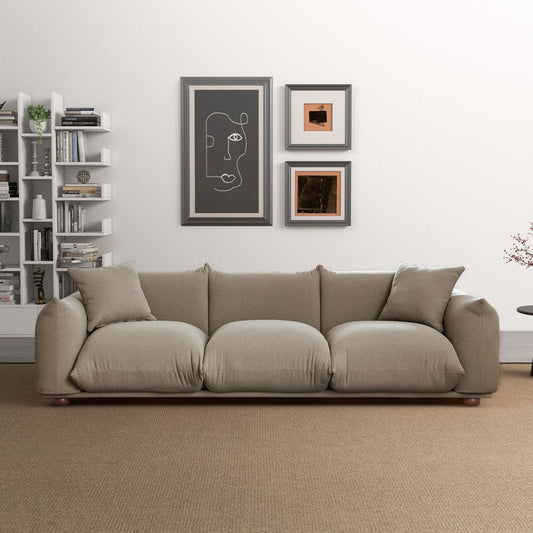 A modern living room with an Ashcroft Furniture Kely Boho Chic Fabric Sofa 100” in light beige, two cushions, a white bookshelf of books and decor, three abstract art frames above the sofa, and a small round side table on the right.
