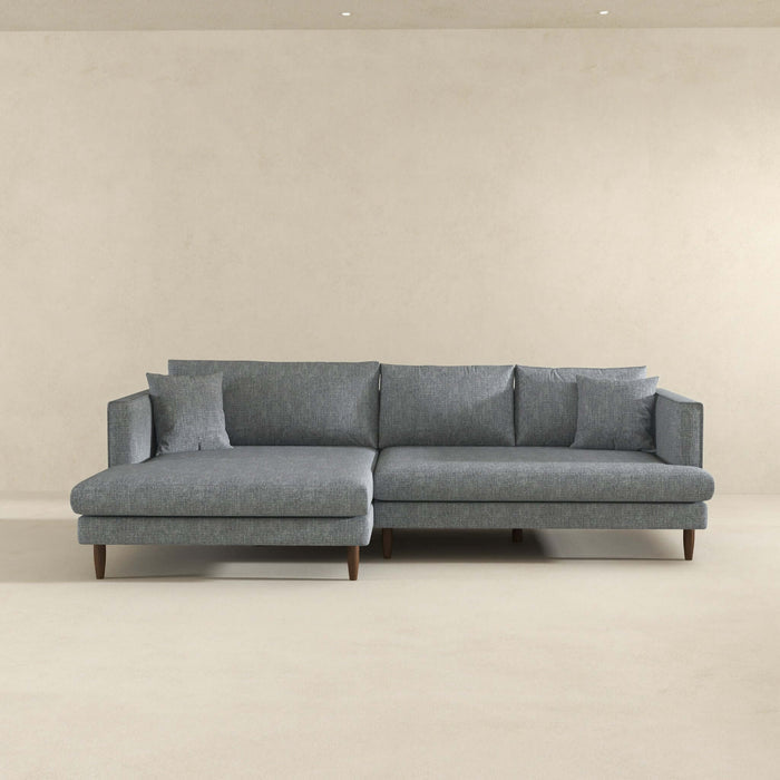 The Ashcroft Furniture Blake Velvet Upholstered L-Shape Chaise Sofa Sectional 107 features plush velvet, wooden legs, and matching cushions, arranged against a beige wall in a minimalist room.