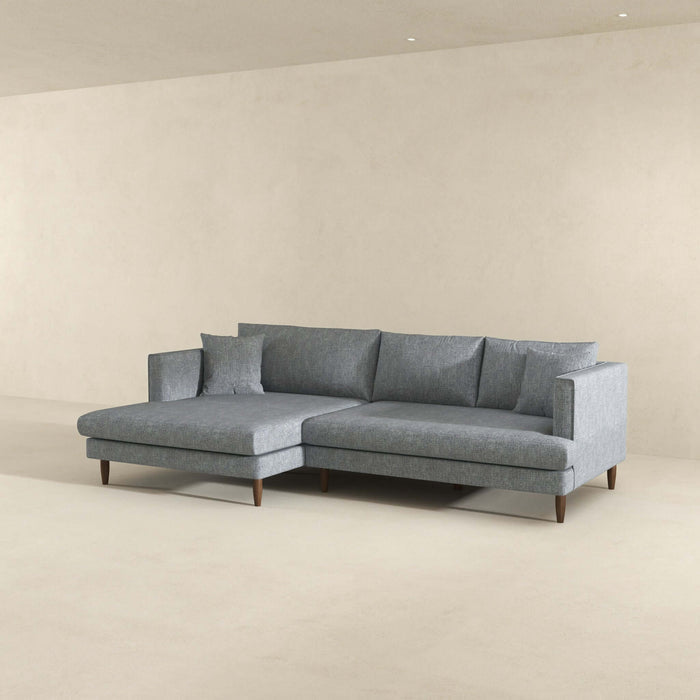The Ashcroft Furniture Blake Velvet Upholstered L-Shape Chaise Sofa Sectional 107 with wooden legs and cushions sits in a minimal empty room, its reversible chaise set against a plain beige wall and floor.