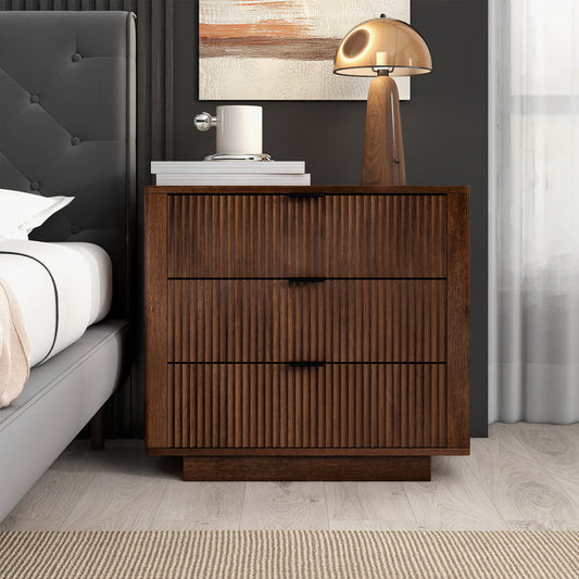 An Ashcroft Furniture Lola Styled Solid Wood Walnut Nightstand 31 (3 Drawers) sits next to a gray upholstered bed, topped with a white cylinder lamp, stacked books, and a gold dome lamp, accented by abstract art and a beige rug.