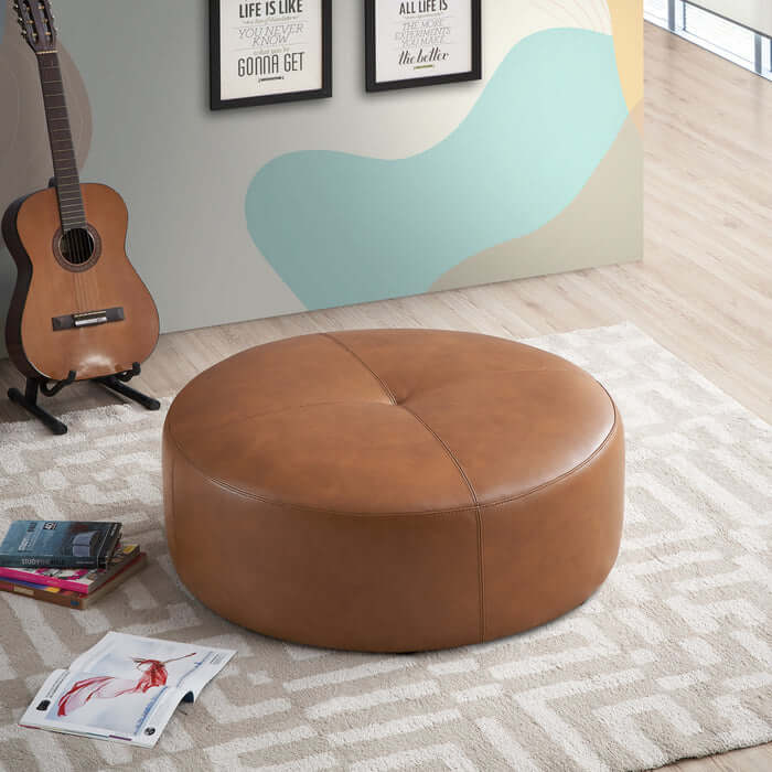 The Ashcroft Furniture Co. Rochelin Round Tan Leather Ottoman 36 features genuine leather and stands on a patterned beige rug in a modern room with a guitar, wall art, and magazines, highlighting subtle mid-century modern style.