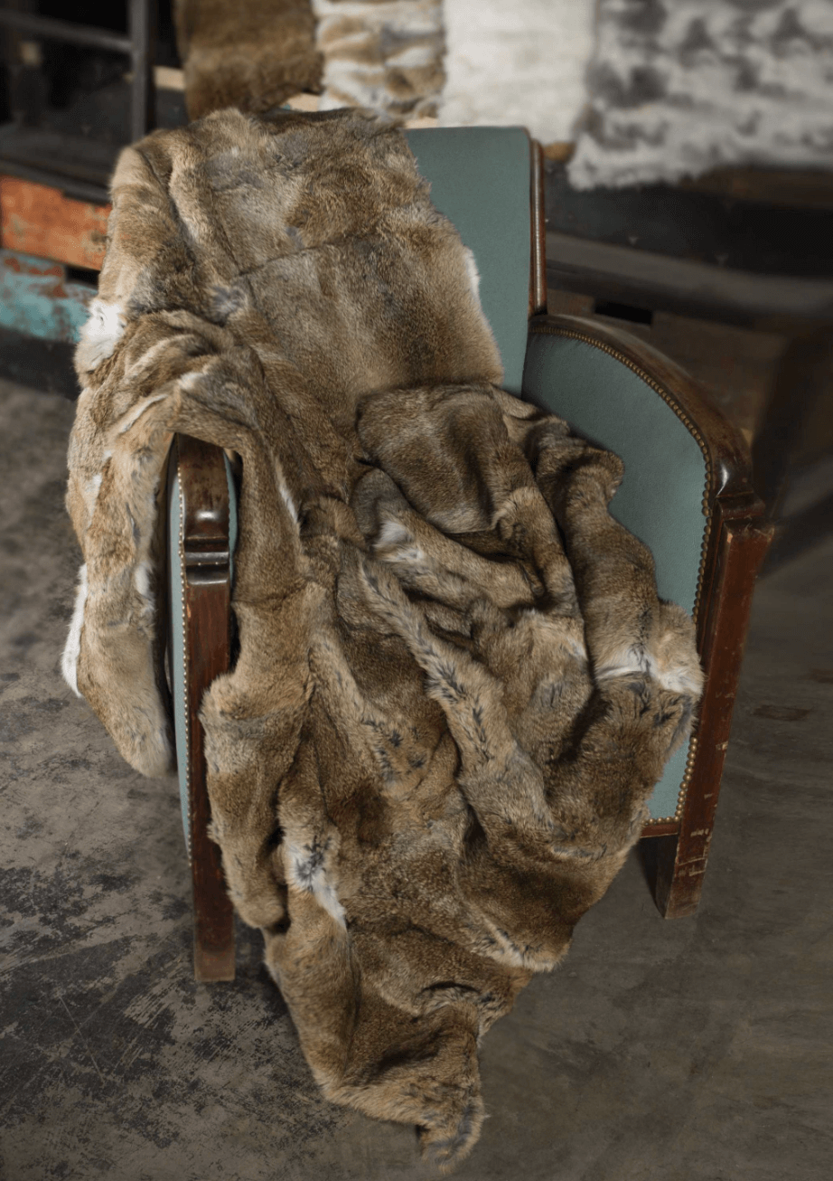 The Home Roots Gray Rabbit Fur Throw Blanket is draped over a vintage wooden chair with teal upholstery, placed on a stone floor in a cozy, dimly lit room.