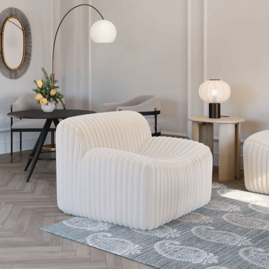 A modern living room featuring the ZUO Modern Osterbro Channel Tufted Cream Faux Fir Armless Lounge Chair on a patterned rug, a round side table with a lamp, a black table with flowers, a wall mirror, and soft lighting.