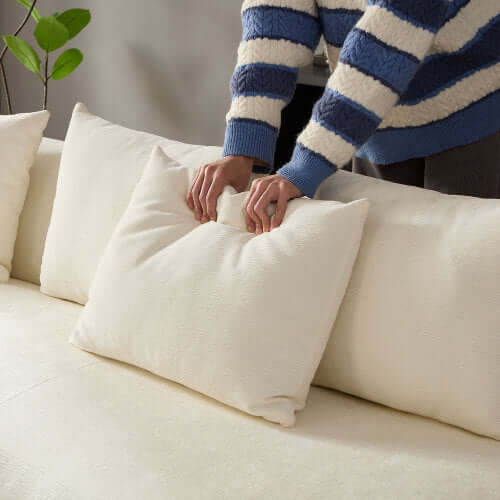Person arranging a plush white pillow on a creamy sofa with soft fabric, showcasing comfort and style.