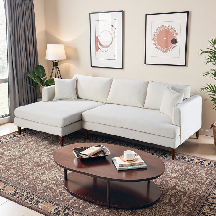 A modern living room features the Ashcroft Furniture Blake Velvet Upholstered L-Shape Chaise Sofa Sectional 107 in white, paired with a dark wood oval coffee table, abstract wall art, a floor lamp, decorative books, and plants on a patterned rug.