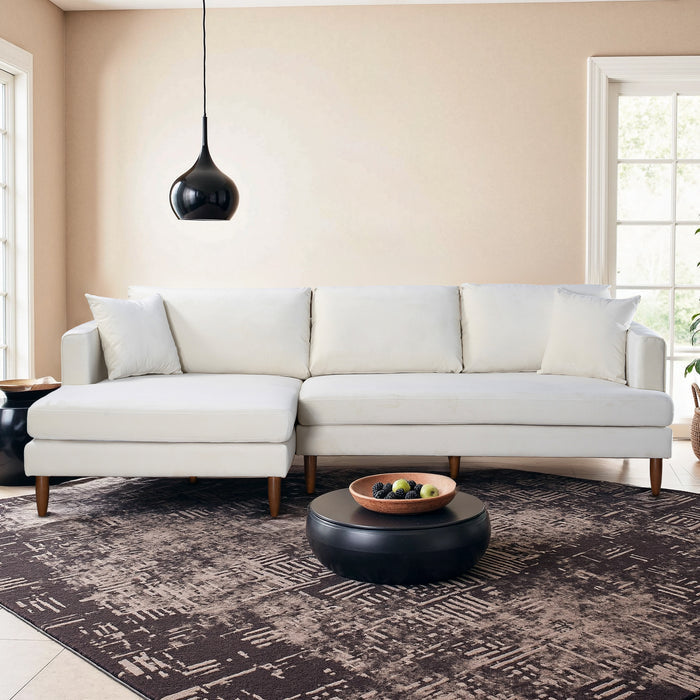 A modern living room with the Ashcroft Furniture Blake Velvet Upholstered L-Shape Chaise Sofa Sectional 107, plush velvet upholstery, a black round coffee table with fruit bowl, patterned rug, black pendant light, and large windows.