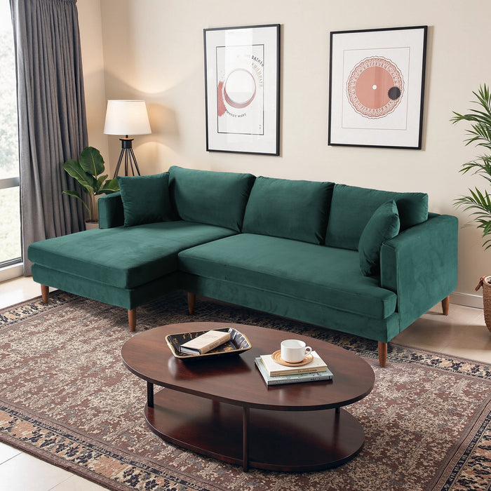 A modern living room with an Ashcroft Furniture Blake Velvet Upholstered L-Shape Chaise Sofa Sectional 107 in green, a round wooden coffee table, patterned rug, framed artwork, potted plant, floor lamp, and gray curtains by the window.