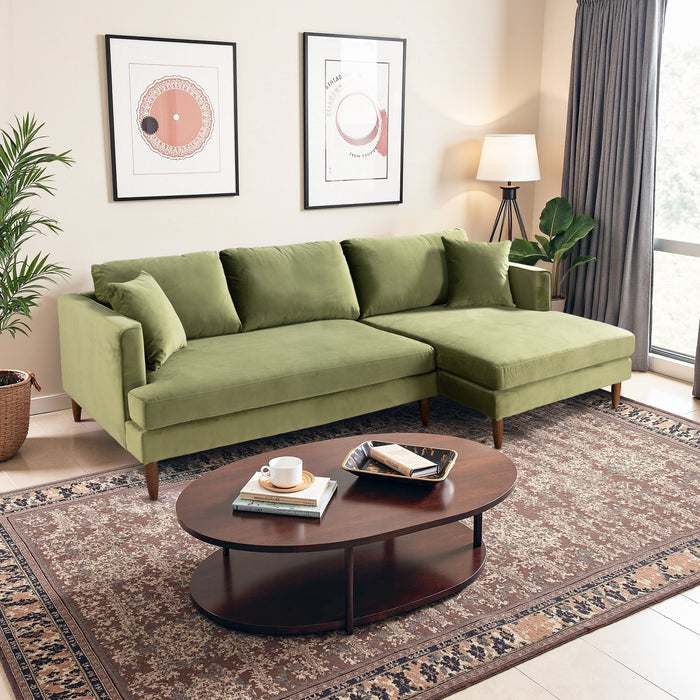 A living room with the Ashcroft Furniture Blake Velvet Upholstered L-Shape Chaise Sofa Sectional 107, four cushions, a round wooden coffee table with books and decor, two framed wall art pieces, a patterned rug, plants, a floor lamp, and window curtains.