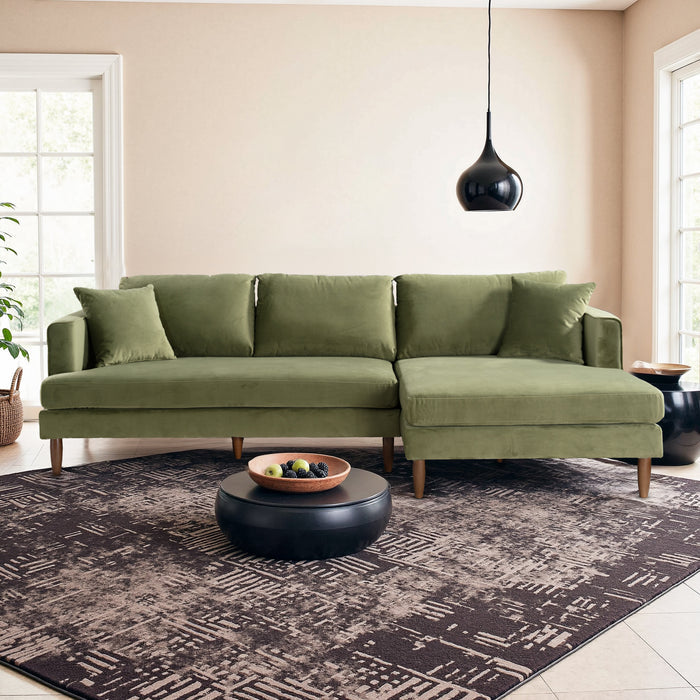A modern living room featuring the Ashcroft Furniture Blake Velvet Upholstered L-Shape Chaise Sofa Sectional 107 in green, two throw pillows, a black round coffee table with a wooden fruit tray, patterned rug, black pendant light, and large windows for natural light.