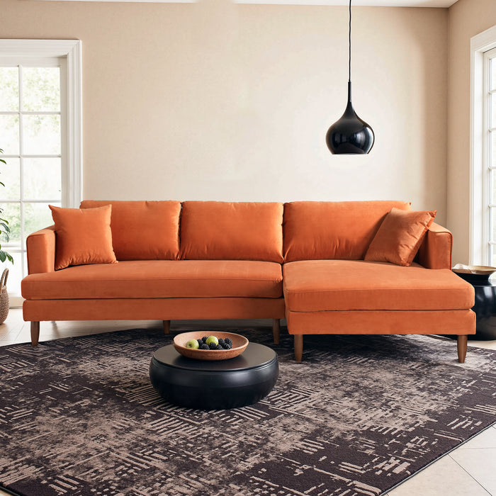 A modern living room showcases the Ashcroft Furniture Blake Velvet Upholstered L-Shape Chaise Sofa Sectional 107 with two matching cushions, a round black coffee table, a geometric rug, and pendant lighting amid large windows.