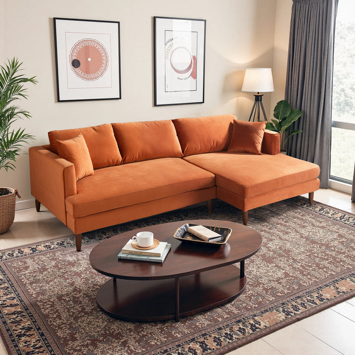 A modern living room with the Ashcroft Furniture Blake Velvet Upholstered L-Shape Chaise Sofa Sectional 107, a round wooden coffee table, framed wall art, standing lamp, potted plants, patterned rug, and large windows with gray curtains.