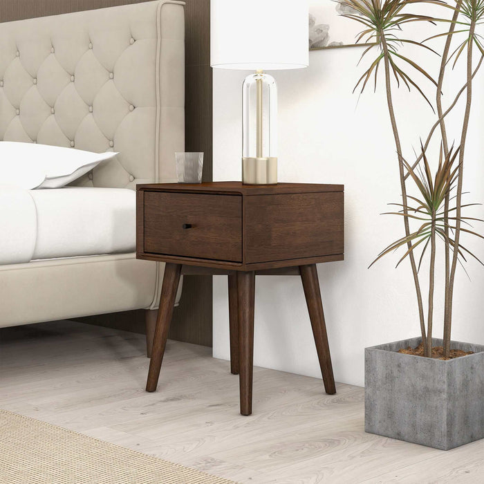 The Avery Solid Wood Nightstand with Drawer by Ashcroft Furniture Co. sits beside a beige tufted bed, topped with a clear glass lamp, while a tall potted plant stands on the light wood floor nearby.