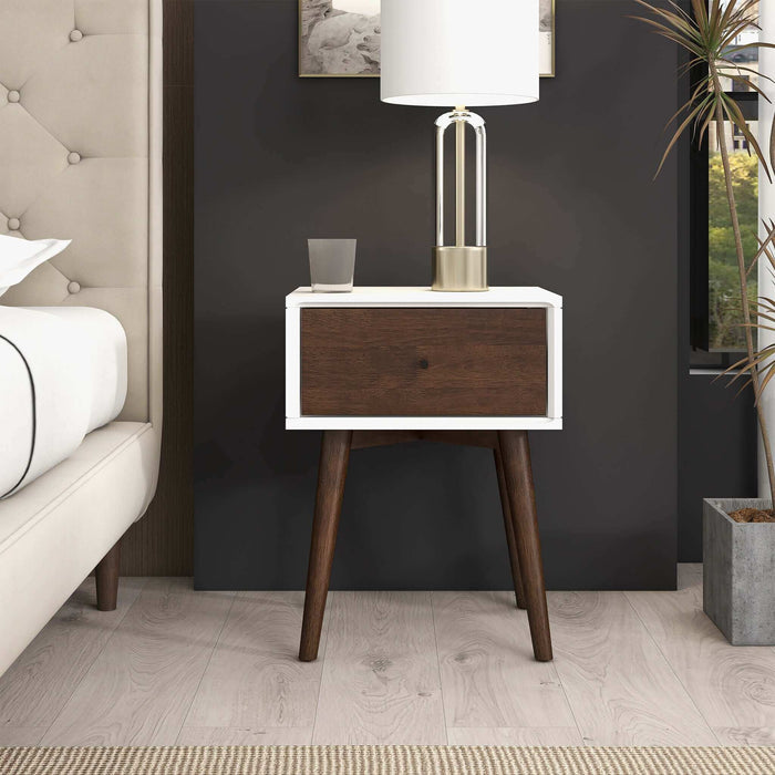 The Avery Solid Wood Nightstand with Drawer by Ashcroft Furniture Co. stands by the bed, showcasing a white frame, walnut finish drawer, and angled legs. A lamp, glass of water, and framed art sit on top; light wood flooring and a rug complete the scene.