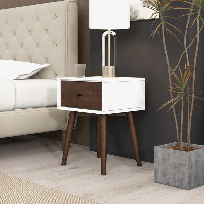 A modern bedroom with a tufted beige headboard, Avery Solid Wood Nightstand with Drawer by Ashcroft Furniture Co., lamp, glass of water, and a tall potted plant in a square gray planter on light wood flooring.