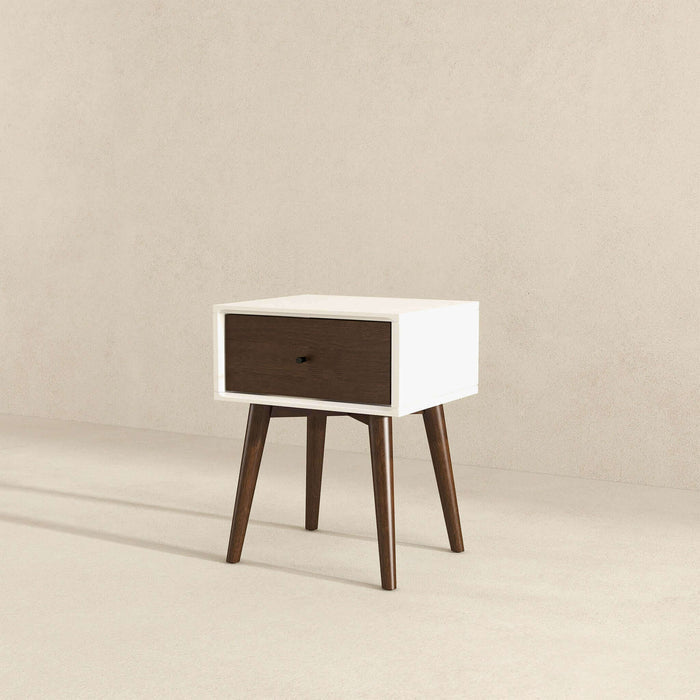 The Avery Solid Wood Nightstand with Drawer by Ashcroft Furniture Co. features a white frame, a walnut-finished dark wood drawer, and four angled dark wood legs, shown against a plain beige wall and floor background.