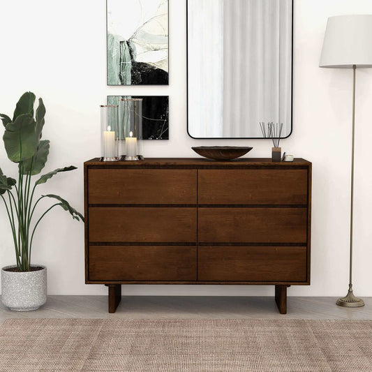 A modern room featuring the Ashcroft Furniture Dubrovnik Walnut Wood Six-Drawer Storage Dresser (47) topped with decor, a plant, and candles. A mirror and abstract art hang above, with a floor lamp on the right and a rug below.