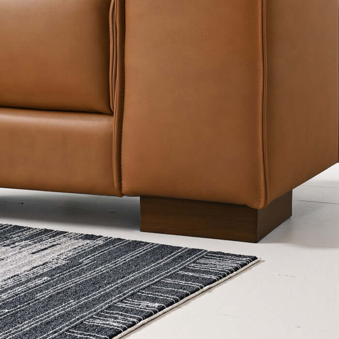 Close-up of the bottom corner of the Ashcroft Furniture Co. Muncie MCM L-Shaped Left-Facing Chaise Sofa Sectional 98W with wooden legs and tan vegan leather, next to a dark blue and white striped rug on a white floor.