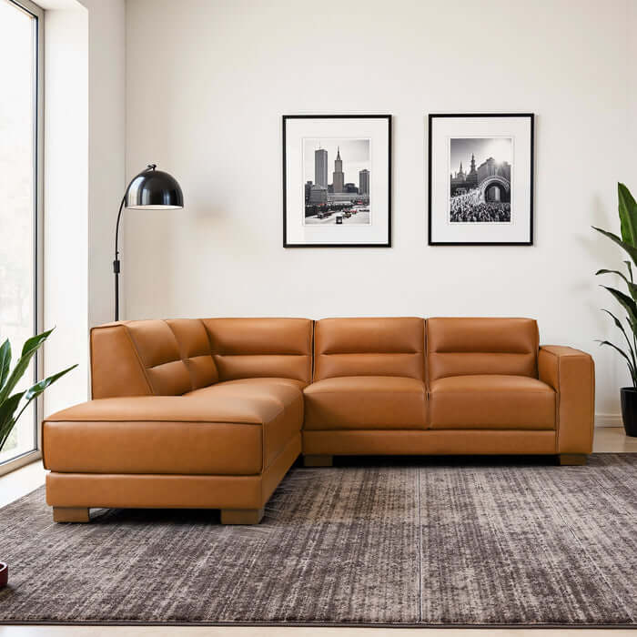 A modern living room features the Ashcroft Furniture Co. Muncie MCM L-Shaped Left-Facing Chaise Sofa Sectional (98W) in tan vegan leather, a gray area rug, floor lamp, potted plants, two cityscape photos, and large windows allowing natural light.