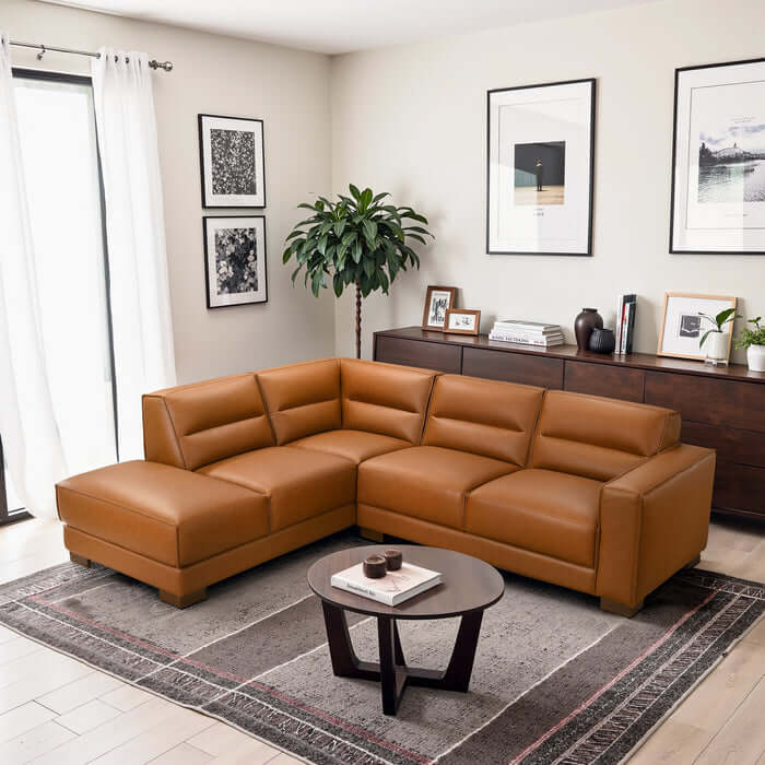 A modern living room featuring the Ashcroft Furniture Co. Muncie MCM L-Shaped Left-Facing Chaise Sofa Sectional (98W) in brown vegan leather, a round coffee table, patterned rug, wall art, sideboard with decor, potted plant, and white curtains.