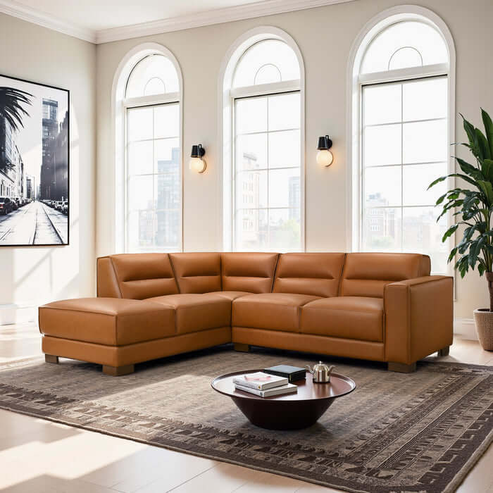 A modern living room features the Ashcroft Furniture Co. Muncie MCM L-Shaped Left-Facing Chaise Sofa Sectional in tan vegan leather, a round coffee table, large patterned rug, potted plant, and black-and-white cityscape photo by tall arched windows.