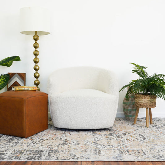 The Mina Cream White Boucle Barrel Swivel Chair by Ashcroft Furniture Co sits on a patterned rug, paired with a brown leather ottoman, tall brass lamp, potted green plants, and decorative books against a white wall.