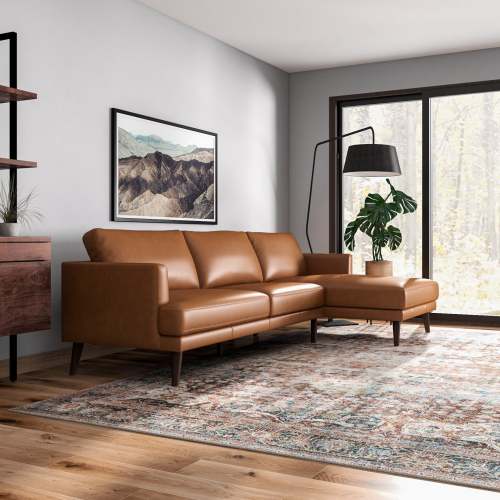 A modern living room with the Ashcroft Furniture Lore MCM Leather L-Shape Chaise Sofa Sectional 105, a patterned rug, large forest-view window, floor lamp, plant, wooden shelf, and a mountain landscape picture on the wall.