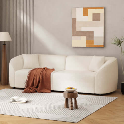 Contemporary sloped arm boucle upholstered sofa in white with brown throw, art, and plants in modern living room setting.